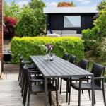 Terrasse mit langem Tisch und Stühlen, umgeben von grünen Sträuchern und einem Gartenhaus.