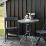 Rundtisch mit zwei Stühlen, Flasche, Gläser und Blumen vor Holzwand.
