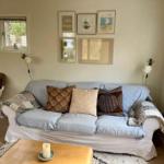 Ein Wohnzimmer mit Sofa, Kissen, Tisch und Fenstern. An der Wand hängen Bilder.