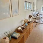 Wohnzimmer mit Holzmöbeln, Wandbildern und Fenstern. Holzdielenboden und Sitzgelegenheiten.