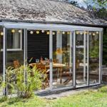 Glass conservatory with wooden table and chairs, string lights, and garden in background.