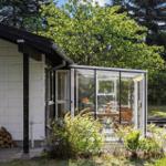 Patio with glass conservatory, wooden table and chairs. Backyard with greenery and trees.