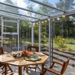 Glass conservatory with wooden table, chairs, and string lights. Outdoor area with greenery and steps.