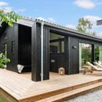 Black wooden house with terrace and loungers. Large windows and covered entrance area.