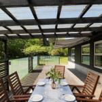 Terrace with table and chairs under a canopy. Background: greenery and house wall.