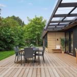 Wooden terrace with seating and grill. Background: green garden and house with glass roof.
