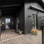 Black wooden house with terrace, grill, and plants. Entrance door and windows visible.