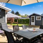 Deck with table, chairs, and umbrella. Small house and green garden in background.
