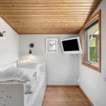 A bedroom with a white bed, wooden floor and ceiling. A TV is mounted on the wall.