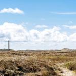 A lighthouse stands atop a dune under a blue sky with white clouds.