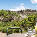 Outdoor area with table and chair, view of dunes and greenery under blue sky.