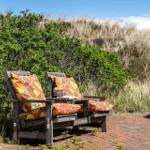 Two wooden chairs with colorful cushions stand on a brick path before grass and bushes.