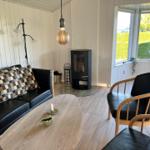 Wohnzimmer mit Sofa, Stuhl, Kamin und Tisch. Durch Fenster Blick auf Grünfläche.
