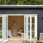 Schlafzimmer mit Bett, Stuhl und Stehlampe. Außen: Holzstühle und Tisch vor einem schwarzen Holzhaus.