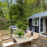 Terrasse mit Grill, Tisch und Stühlen. Im Hintergrund Wald und Ferienhaus mit offener Tür.