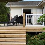 Deck with umbrella, lounge chairs, and table. Wooden steps lead to the deck. Background: house and garden.
