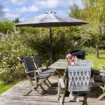 Deck with table, chairs, umbrella, and grill. Background: green garden with shrubs.