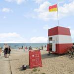 Beach with lifeguard station, flag, and sign. People stroll and swim.