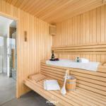Sauna mit Holzbänken, Handtuch, Eimer und Flasche. Tür führt zu einem Bad.