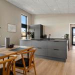 Modern kitchen with dining area and large windows. Wooden floor and gray cabinets.