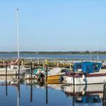 Boats are moored at a wooden pier in the water. In the background, fields and a clear sky are visible.