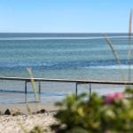 A red kayak floats on the water. A pier leads to the beach. Grass and flowers are in the foreground.