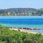 Strand mit klarem Wasser und grüner Vegetation im Vordergrund. Im Hintergrund sind Häuser und Bäume zu sehen.