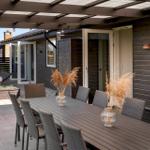Terrasse mit Esstisch, Stühlen und Sonnenschirm. Holzwände und Glastüren führen ins Haus.