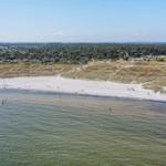 Strand mit Sand und Wasser, dahinter Hügel mit Bäumen und Häusern.