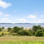 Wind turbine by water with green areas and trees in foreground.