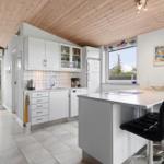 Kitchen with white cabinets, kitchen island, and black bar stools. Wooden ceiling and window with view outside.