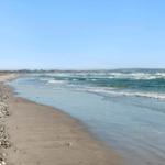 Strand mit Kies und Sand, Wellen brechen am Ufer, Horizont mit Hügeln im Hintergrund.