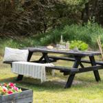 Holzpicknicktisch mit Decke und Kissen im Garten. Hintergrund: Gras und Bäume.