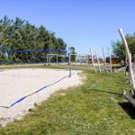 Sandplatz mit Volleyballnetz, Holzspielplatz und Picknicktisch im Gras.