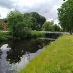 Flussufer mit Brücke, Gras und Bäumen. Im Hintergrund ein rotes Haus.