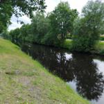 Ein ruhiger Kanal mit grüner Ufervegetation und Bäumen. Ein Haus ist am gegenüberliegenden Ufer sichtbar.