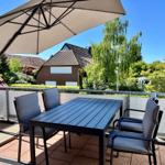 Terrasse mit Tisch, vier Stühlen und Sonnenschirm. Blick auf begrünte Umgebung und Nachbarhäuser.