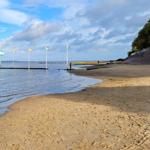 Strand mit Steg und Flaggen im Wasser, Himmel mit Wolken, Bäume am Ufer.