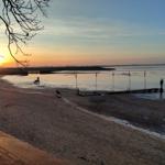 Sonnenuntergang am Strand mit Steg und Windkraftanlagen im Hintergrund.