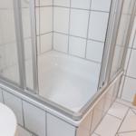 Dusche mit Glaswand und weißen Fliesen. Holztür und weißer Waschtisch sichtbar.