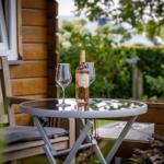 Auf der Terrasse steht ein Tisch mit Weinflasche und zwei Gläsern. Hintergrund: Holzhaus und grüne Pflanzen.