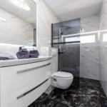 Badezimmer mit Waschbecken, Dusche und Toilette. Marmorwände und dunkler Boden.