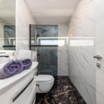 Badezimmer mit Dusche, Waschbecken und Toilette. Marmorwände und dunkler Boden.