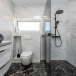 Badezimmer mit Dusche, Toilette und Waschbecken. Weiße Marmorwände, schwarzer Boden und Fenster.