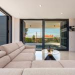 Ein Wohnbereich mit Sofa, Tisch und Fernseher. Durchgehende Glaswände öffnen sich zu einer Poolterrasse.