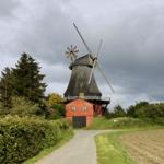 Alte Windmühle mit rotem Unterbau und grauem Dach, umgeben von Wald und Feldern.