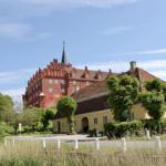 Rotes Schloss mit Turm und gelbem Nebengebäude unter blauem Himmel.