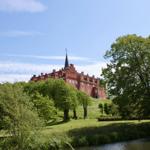 Rotes Schloss auf Hügel mit Grünflächen und Fluss im Vordergrund.