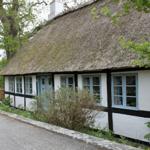 Haus mit Strohdach, weißen Wänden und blauen Fenstern. Umgeben von Bäumen und Pflanzen.