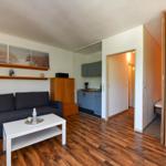 Wohnzimmer mit Sofa, Couchtisch und Küchenbereich. Holzfußboden und Schlafzimmer sichtbar.